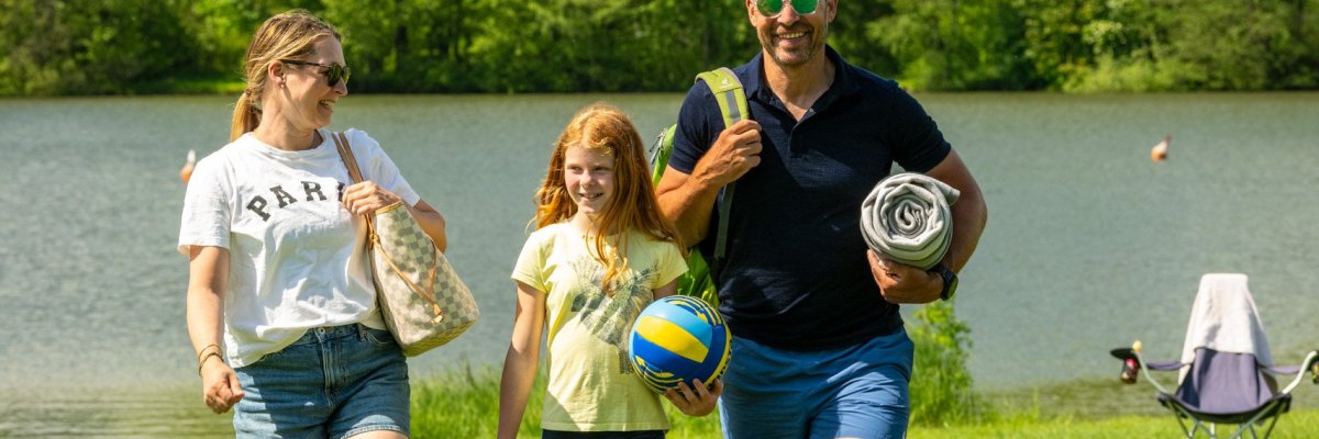 Eine fröhliche Familie spielt am Ufer des Secker Weihers in Rennerod im Hohen Westerwald Ball; im Hintergrund spiegeln sich Bäume im ruhigen Wasser, und eine weitläufige Wiese lädt zum Verweilen ein.