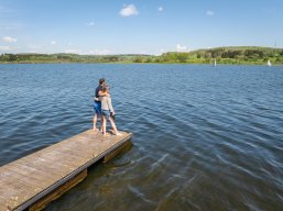 Die Krombachtalsperre bei Rennerod im Hohen Westerwald ist eine große Seelandschaft. Entlang des Ufers führen gut ausgebaute Wanderwege, die zu entspannten Spaziergängen einladen, während auf dem Wasser zahlreiche Freizeitmöglichkeiten wie Segeln, Bootfahren oder Surfen auf ihren Genuss warten. Auf einem Holzsteg gehen zwei Personen barfuß, genießen die frische Luft und lassen ihre Füße sanft das kühle Wasser berühren. Diese idyllische Szenerie bietet sowohl Einheimischen als auch Besuchern den idealen Ort, um abzuschalten und die Natur in vollen Zügen zu erleben. Dieses Ausflugsziel überzeugt durch die Größe und den Westerwaldwind und weitere abwechslungsreiche Freizeitangebote. Die Krombachtalsperre ist das Segel- und Surf-Paradies im Westerwald bei Rennerod im Hohen Westerwald. Wassersport mit Segelboot, Tretboot, SUP und Surfbrett möglich. Auch Barfussbaden ist möglich. Hier geniessen 2 Wanderer dieses Erlebnis.